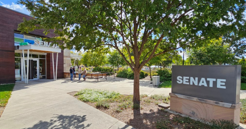 A modern building with a sign reading "SENATE," surrounded by greenery and people outside.