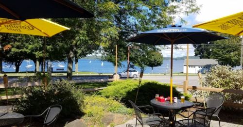 A scenic view of a lakeside patio with umbrellas, tables, and greenery, overlooking the water and distant sailboats.
