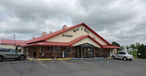 A bakery with a red roof and large windows, featuring outdoor seating and a parking lot under a cloudy sky.