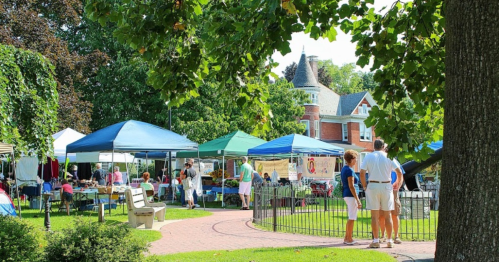 A vibrant outdoor market with colorful tents, people browsing, and a historic building in the background surrounded by greenery.