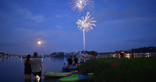 People watch fireworks over a lake while standing near colorful kayaks at dusk.
