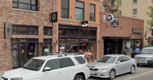 A street view of brick buildings with shops, parked cars, and people sitting outside on a sunny day.
