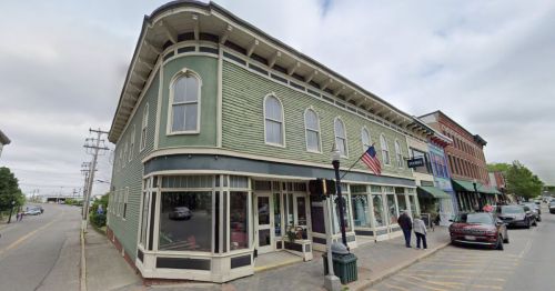 Historic green building on a street corner, featuring large windows and an American flag, with shops nearby.