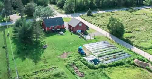 Aerial view of a red house with a garden and tractor, surrounded by green fields and a road nearby.