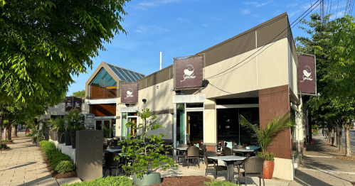A modern restaurant exterior with outdoor seating, large windows, and greenery on a sunny day.