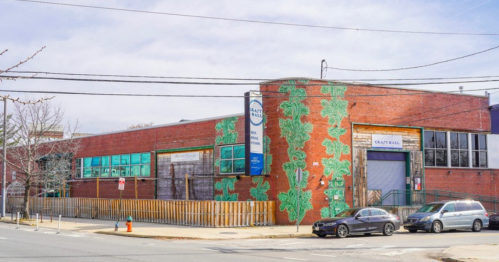 A brick building with green leaf murals, featuring large windows and a fenced area, set against a cloudy sky.