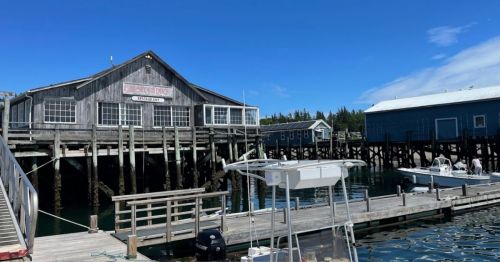 A wooden building on stilts by the water, with boats docked nearby and a clear blue sky above.