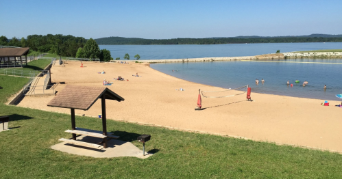 A sandy beach by a calm lake, with picnic areas, volleyball nets, and people enjoying the water on a sunny day.