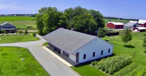 Aerial view of a white farmhouse surrounded by green fields, trees, and red barns under a clear blue sky.