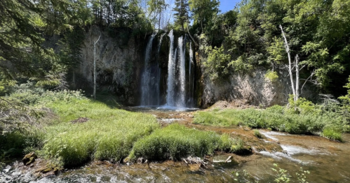 A serene waterfall cascades over rocky cliffs, surrounded by lush greenery and a clear stream below.