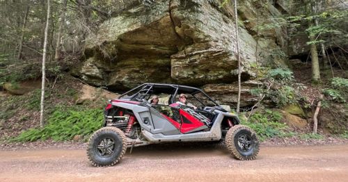 A person sits in a black and red off-road vehicle parked on a dirt road near a large rock formation in a forested area.