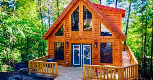 A cozy wooden cabin with large windows, surrounded by trees and featuring a spacious deck.