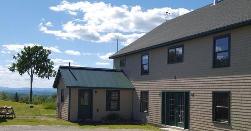 A two-story house with a green roof, surrounded by grass and trees, under a blue sky with fluffy clouds.