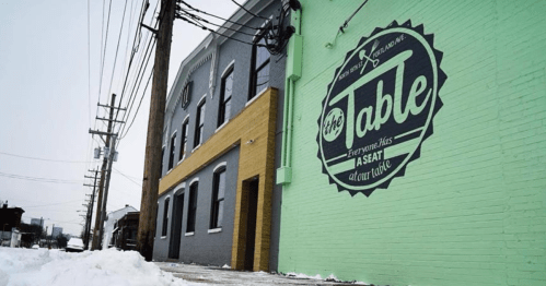 A colorful mural on a building reads "the Table: Everyone Has a Seat at Our Table," with snow on the ground.