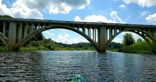 A view from a kayak on a river, featuring a large arched bridge under a blue sky with fluffy clouds.