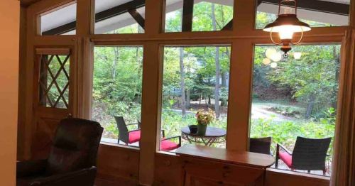 Cozy interior view with large windows overlooking a green, wooded area and outdoor seating.