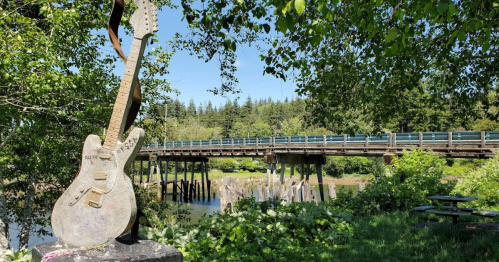 A guitar sculpture in the foreground with a wooden bridge and lush greenery in the background.