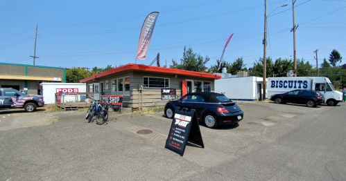A small shop with a red roof, a black convertible car parked outside, and food trucks nearby on a sunny day.