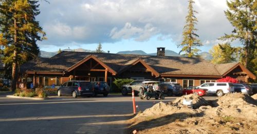 A rustic lodge with a wooden exterior, surrounded by trees and mountains, with parked cars and construction nearby.