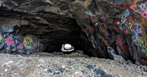 A rocky cave entrance with colorful graffiti on the walls and a view of light at the far end.