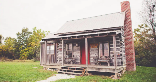 A rustic log cabin with a red door, surrounded by greenery and featuring a porch with wooden chairs.