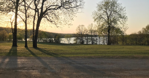 A serene lakeside view at sunset, with trees casting long shadows on a grassy area.