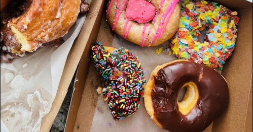 A box of colorful donuts, including a chocolate frosted, a sprinkle-covered, and a fruity cereal-topped donut.