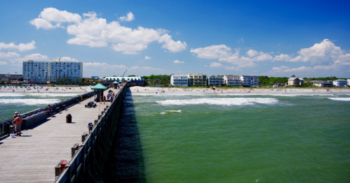 A wooden pier extends into the ocean, with beachgoers and buildings visible along the shore under a blue sky.