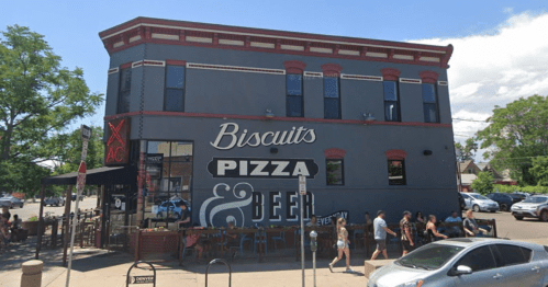 A gray building with a large sign reading "Biscuits Pizza & Beer," bustling with people outside on a sunny day.