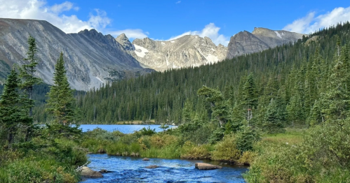 A serene landscape featuring a clear stream, lush greenery, and majestic mountains under a blue sky with scattered clouds.