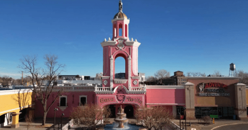 A colorful pink building with a tall tower and fountain, featuring a whimsical design, set against a clear blue sky.