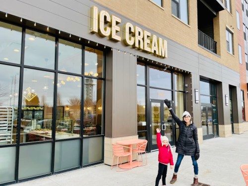 A woman and a child joyfully pose outside an ice cream shop, waving with a sign that reads "ICE CREAM" above them.