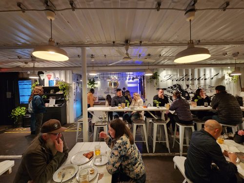 A lively indoor dining space with people enjoying meals at tables, plants, and a modern decor.