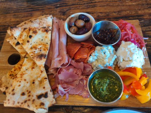 A wooden platter with assorted meats, cheeses, olives, pickled vegetables, and flatbread.