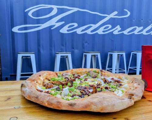 A delicious pizza topped with green onions and meat, set on a wooden table with a blue background and bar stools.