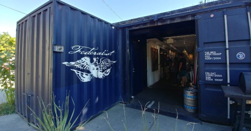 Entrance of a shipping container bar named "Federalist," with people inside and a keg outside.