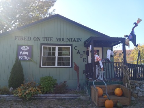 A green café building with a sign reading "Fired on the Mountain," decorated with pumpkins and autumn foliage.