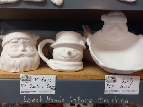 Three ceramic holiday decorations: a Santa mug, a snowman mug, and a Santa bowl, displayed on a shelf with price tags.