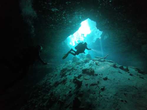 Two divers explore an underwater cave, illuminated by sunlight filtering through an opening above.