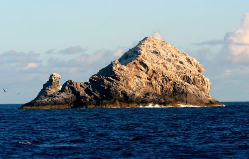 A rocky island rises from the ocean, surrounded by calm blue waters and a clear sky.