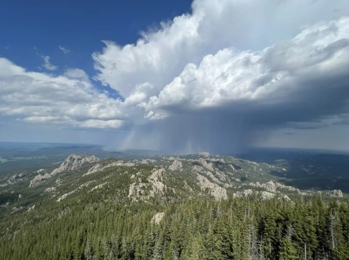A panoramic view of rugged mountains under a dramatic sky with clouds and distant rain. Lush green forests in the foreground.