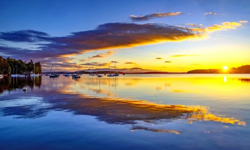 A serene lake at sunset, with colorful clouds reflecting on the water and boats anchored peacefully.