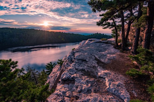 A rocky cliff overlooks a tranquil lake at sunset, surrounded by trees and a colorful sky.