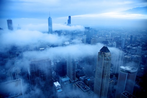 Aerial view of a city skyline shrouded in fog, with tall buildings emerging through the clouds under a blue sky.