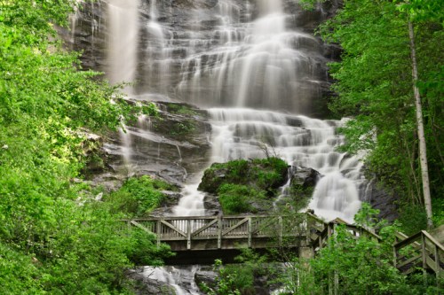 A serene waterfall cascades down rocky cliffs, surrounded by lush green trees and a wooden bridge in the foreground.