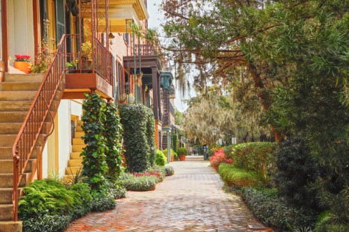 A charming brick pathway lined with lush greenery and colorful flowers, flanked by historic buildings and stairs.