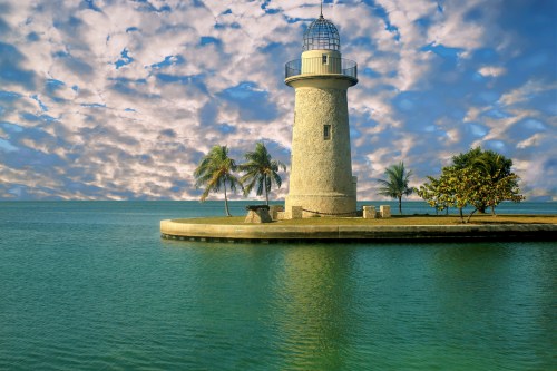 A stone lighthouse stands on a small island, surrounded by calm waters and palm trees under a cloudy sky.