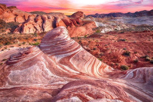 Vibrant rock formations with swirling patterns under a colorful sunset sky in a desert landscape.