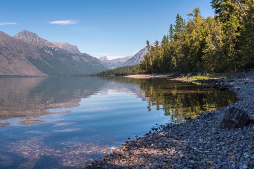 A serene lake surrounded by mountains and trees, with a rocky shoreline reflecting the clear blue sky.