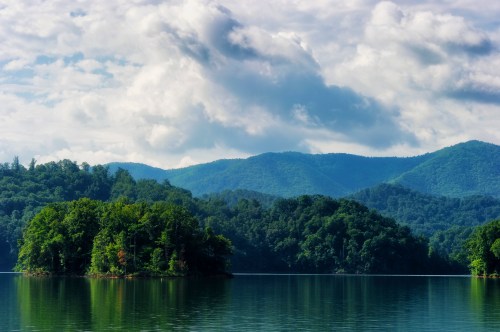 A serene lake surrounded by lush green hills and mountains under a partly cloudy sky.